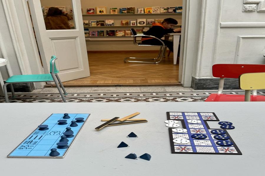 En la imagen se ven los tableros y fichas de los dos juegos sobre una mesa, mientras que de fondo se puede apreciar parte de La Casa Imaginada de la biblioteca