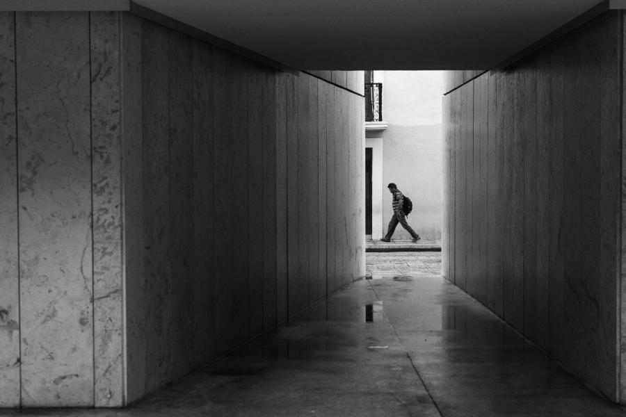 Fotografía en blanco y negro con un pasillo donde se puede ver a una persona caminando con una mochila colgada 